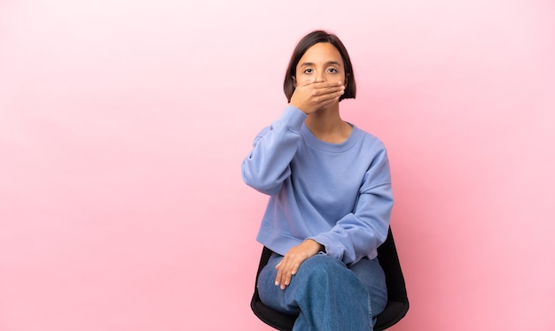 Jeune femme métisse assise sur une chaise isolée sur fond rose couvrant la bouche avec la main