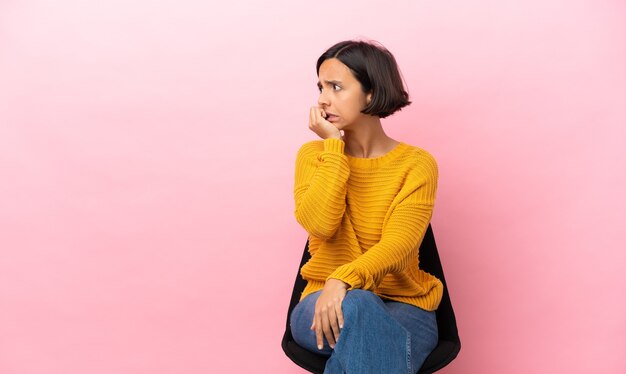 Jeune femme métisse assise sur une chaise isolée sur fond rose est un peu nerveuse