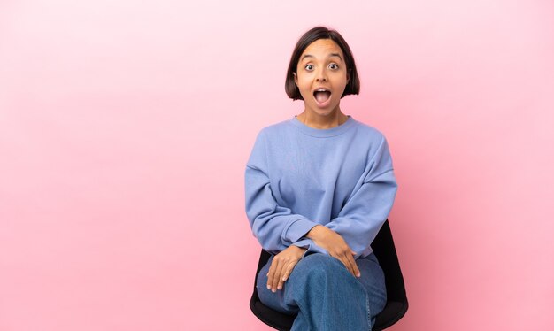 Jeune femme métisse assise sur une chaise isolée sur fond rose avec une expression faciale surprise