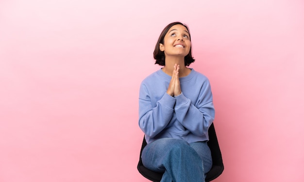 Jeune femme métisse assise sur une chaise isolée sur fond rose maintient la paume ensemble. La personne demande quelque chose