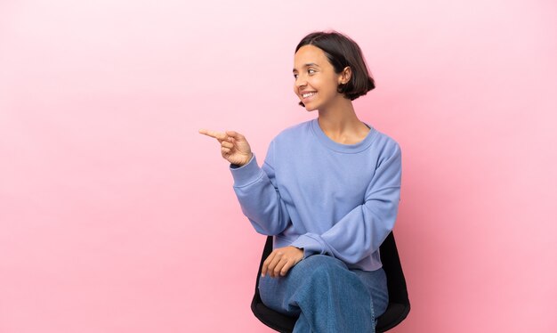 Jeune femme métisse assise sur une chaise isolée sur fond rose, pointant le doigt sur le côté