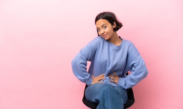 Photo jeune femme métisse assise sur une chaise isolée sur fond rose posant avec les bras à la hanche et souriant