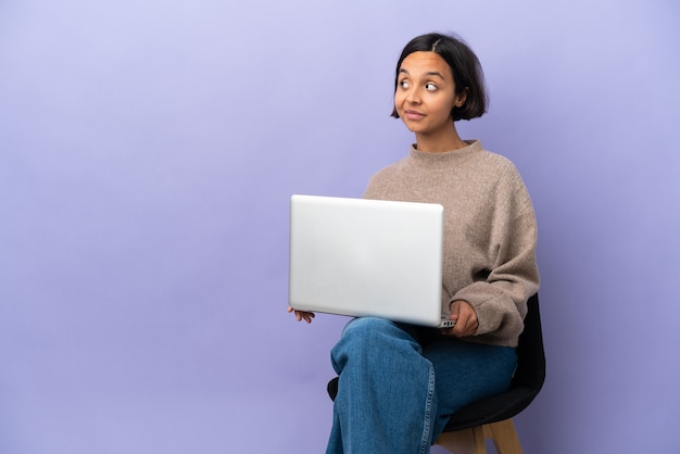 Jeune femme métisse assise sur une chaise avec un ordinateur portable isolé sur fond violet ayant des doutes tout en regardant de côté