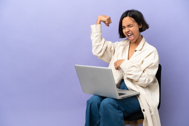 Jeune femme métisse assise sur une chaise avec ordinateur portable isolé sur fond violet célébrant une victoire