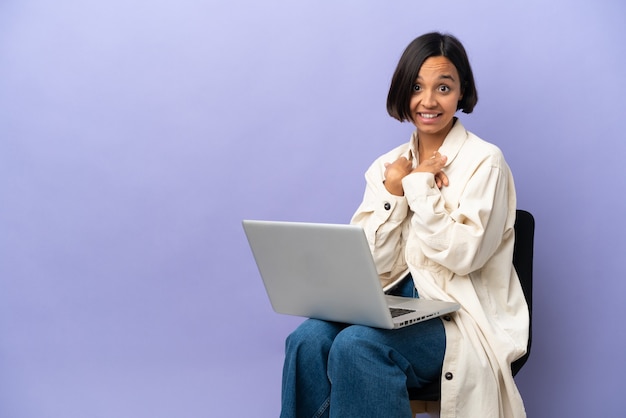 Jeune femme métisse assise sur une chaise avec un ordinateur portable isolé sur fond violet avec une expression faciale surprise