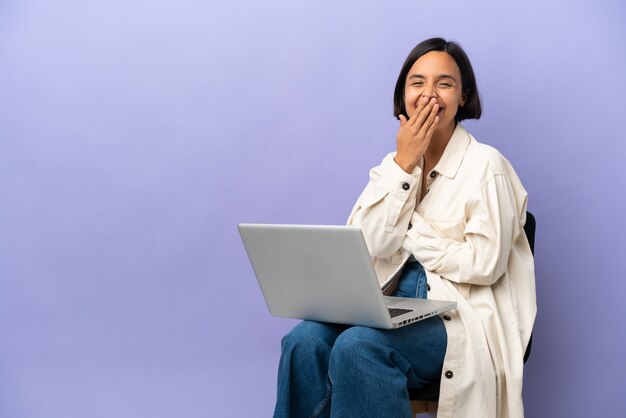 Jeune femme métisse assise sur une chaise avec ordinateur portable isolé sur fond violet heureux et souriant couvrant la bouche avec la main