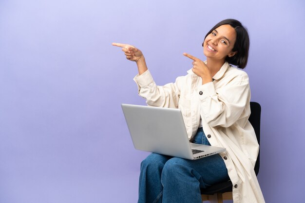 Jeune femme métisse assise sur une chaise avec un ordinateur portable isolé sur fond violet, pointant le doigt sur le côté et présentant un produit