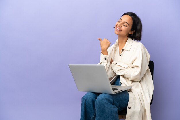 Jeune femme métisse assise sur une chaise avec ordinateur portable isolé sur fond violet pointant vers le côté pour présenter un produit