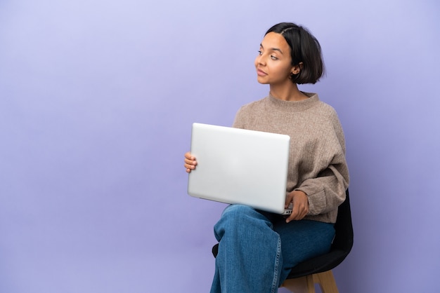 Jeune femme métisse assise sur une chaise avec ordinateur portable isolé sur fond violet en position arrière et à côté