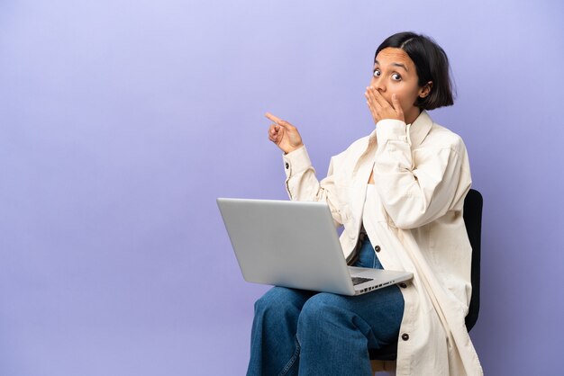 Jeune femme métisse assise sur une chaise avec ordinateur portable isolé