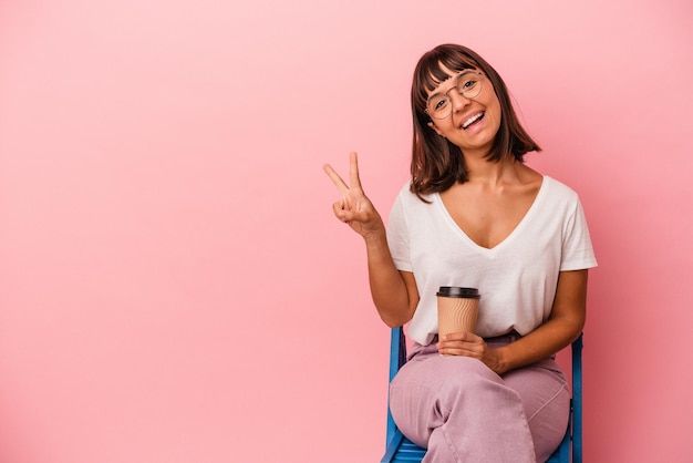 Jeune femme métisse assise sur une chaise tenant un café isolé sur fond rose joyeuse et insouciante montrant un symbole de paix avec les doigts