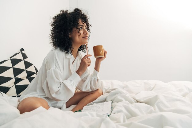 Jeune femme métisse assise sur le lit en prenant un café et en écrivant sur son journal Copier l'espace