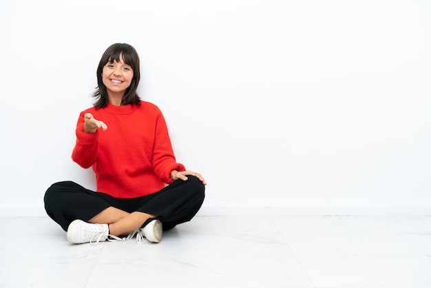 Jeune femme métisse assise sur le sol isolé sur fond blanc se serrant la main pour conclure une bonne affaire