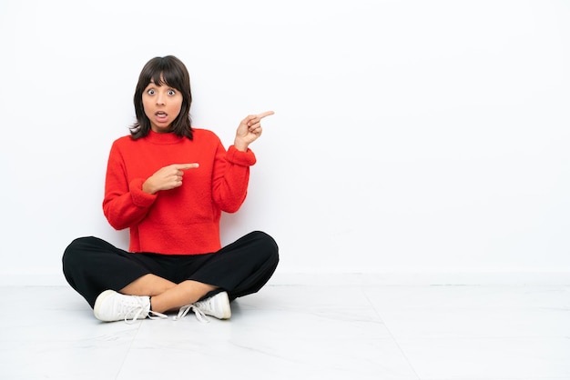 Jeune femme métisse assise sur le sol isolé sur fond blanc surpris et pointant du côté