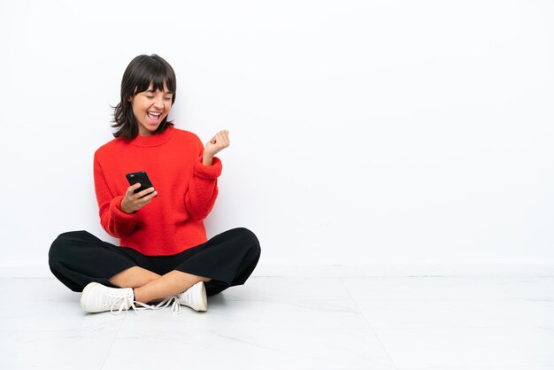 Jeune femme métisse assise sur le sol isolé sur fond blanc avec téléphone en position de victoire