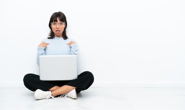 Jeune femme métisse assise sur le sol avec un ordinateur portable isolé sur fond blanc avec une expression faciale surprise