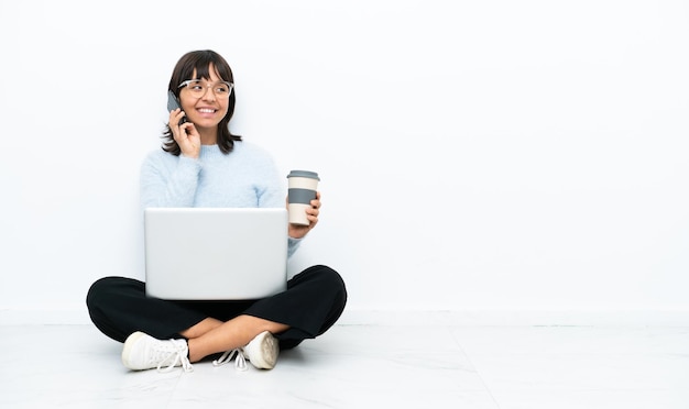 Jeune femme métisse assise sur le sol avec un ordinateur portable isolé sur fond blanc tenant du café à emporter et un mobile