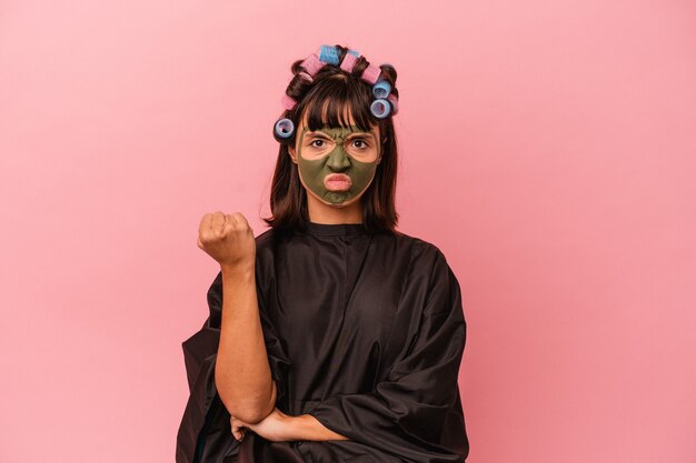 Jeune femme métisse en attente dans un salon Beaty isolé sur fond rose montrant le poing à la caméra, expression faciale agressive.