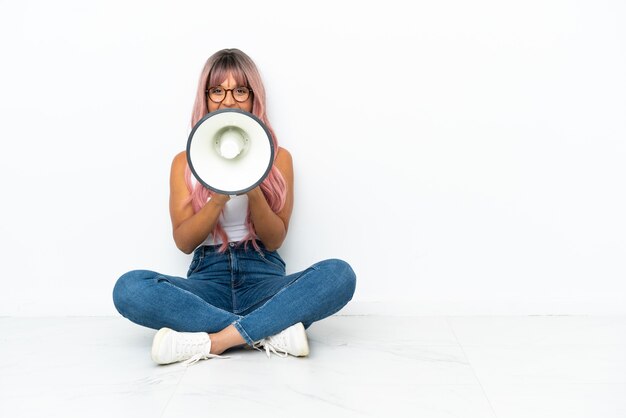 Jeune femme métisse aux cheveux roses assis sur le sol isolé sur fond blanc criant à travers un mégaphone