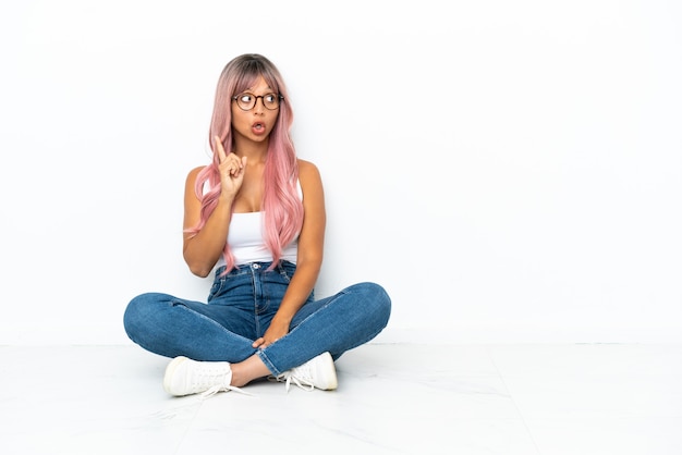 Jeune femme métisse aux cheveux roses assis sur le sol isolé sur fond blanc pensant à une idée pointant le doigt vers le haut