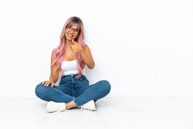 Jeune femme métisse aux cheveux roses assise sur le sol isolée sur fond blanc avec les pouces vers le haut parce que quelque chose de bien s'est produit