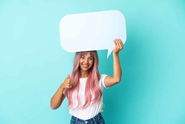 Jeune femme métisse aux cheveux roses isolée sur fond bleu tenant une bulle de dialogue vide avec le pouce vers le haut