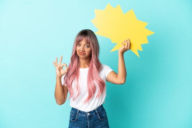 Jeune femme métisse aux cheveux roses isolée sur fond bleu tenant une bulle vide et faisant signe OK