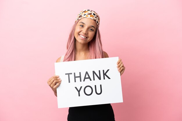 Jeune femme métisse aux cheveux roses isolée sur fond rose tenant une pancarte avec du texte MERCI avec une expression heureuse