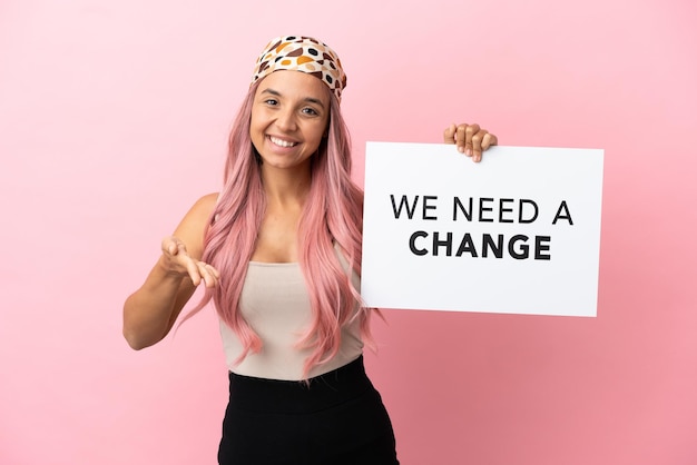 Jeune femme métisse aux cheveux roses isolée sur fond rose tenant une pancarte avec du texte Nous avons besoin d'un changement pour conclure un accord