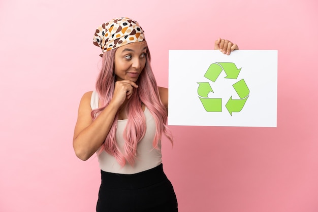 Photo jeune femme métisse aux cheveux roses isolée sur fond rose tenant une pancarte avec l'icône de recyclage