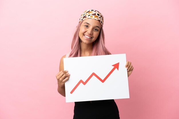 Photo jeune femme métisse aux cheveux roses isolée sur fond rose tenant une pancarte avec un symbole de flèche de statistiques en croissance avec une expression heureuse