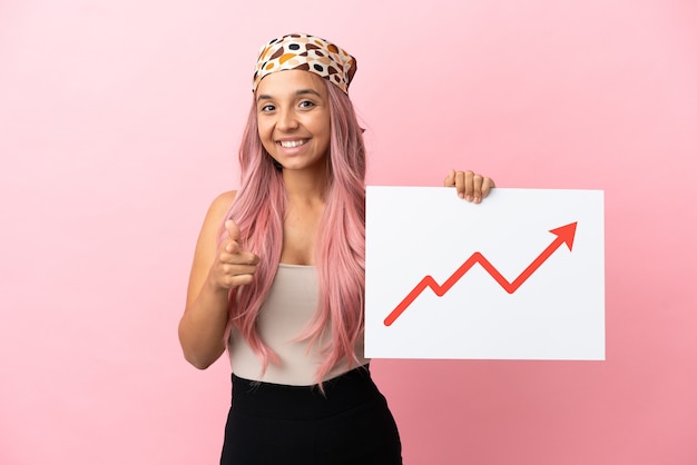 Jeune femme métisse aux cheveux roses isolée sur fond rose tenant une pancarte avec un symbole de flèche de statistiques croissantes et pointant vers l'avant