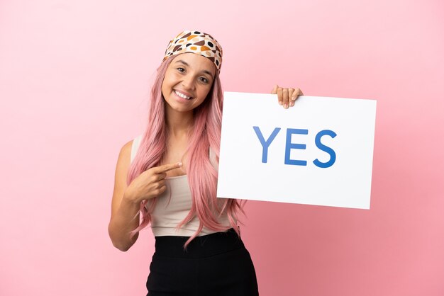 Jeune femme métisse aux cheveux roses isolée sur fond rose tenant une pancarte avec le texte OUI et le pointant