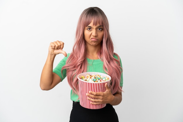 Jeune femme métisse aux cheveux roses mangeant du pop-corn isolé sur fond blanc montrant le pouce vers le bas avec une expression négative