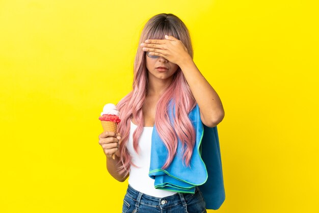 Jeune femme métisse aux cheveux roses tenant une crème glacée isolée sur fond jaune couvrant les yeux à la main. Je ne veux pas voir quelque chose