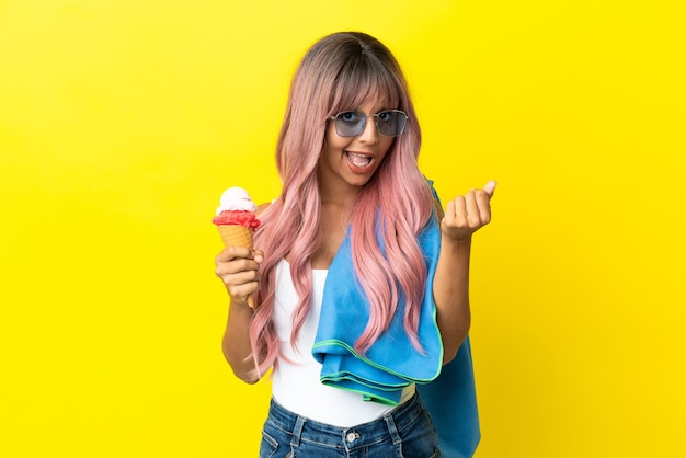 Jeune femme métisse aux cheveux roses tenant une glace isolée sur fond jaune faisant un geste d'argent