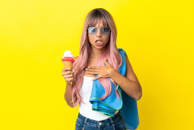 Jeune femme métisse aux cheveux roses tenant une glace isolée sur fond jaune surprise et choquée en regardant à droite