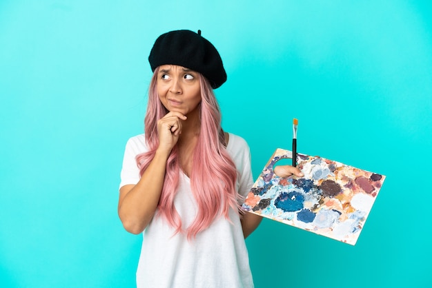 Photo jeune femme métisse aux cheveux roses tenant une palette isolée sur fond bleu ayant des doutes et pensant