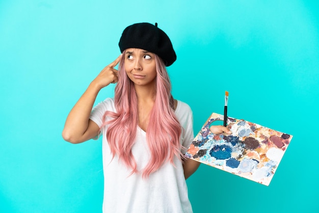 Photo jeune femme métisse aux cheveux roses tenant une palette isolée sur fond bleu ayant des doutes et pensant