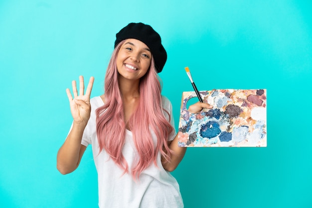 Jeune femme métisse aux cheveux roses tenant une palette isolée sur fond bleu heureux et comptant quatre avec les doigts