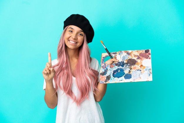 Jeune femme métisse aux cheveux roses tenant une palette isolée sur fond bleu montrant et levant un doigt