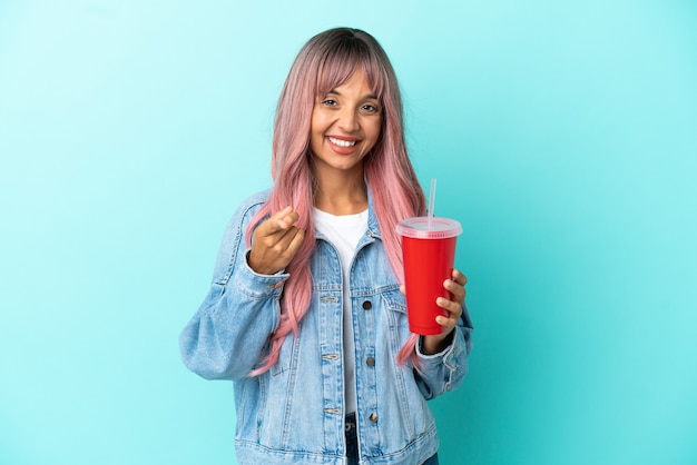 Jeune femme métisse buvant une boisson fraîche isolée sur fond bleu pointant vers l'avant avec une expression heureuse