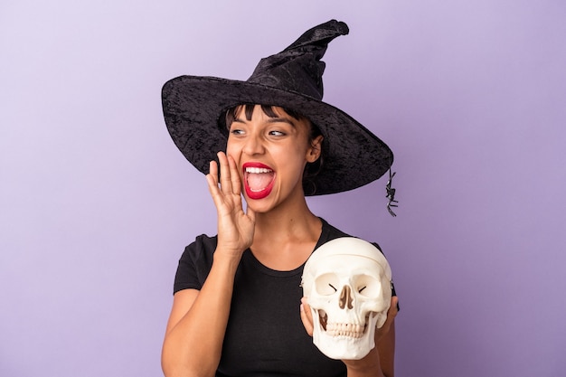 Jeune femme métisse déguisée en sorcière tenant un crâne isolé sur fond violet criant et tenant la paume près de la bouche ouverte.