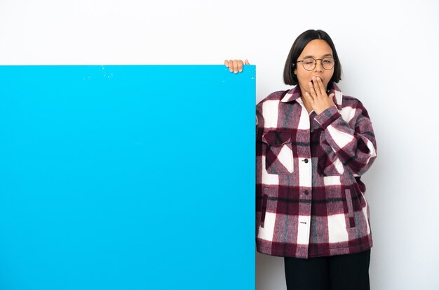 Jeune femme métisse avec une grande pancarte bleue isolée sur fond blanc bâillant et couvrant la bouche grande ouverte avec la main