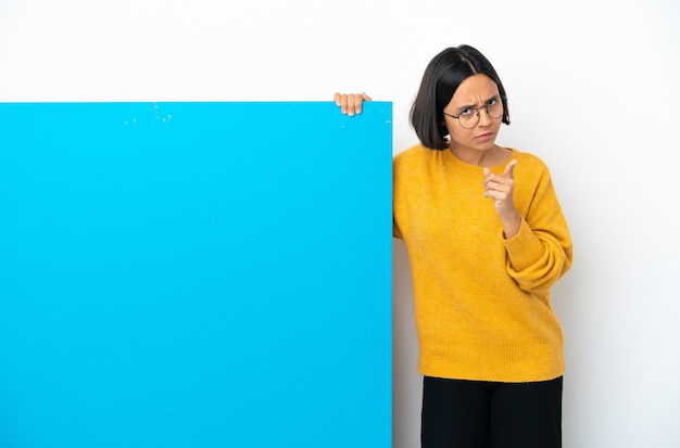 Jeune femme métisse avec une grande pancarte bleue isolée sur fond blanc frustré et pointant vers l'avant