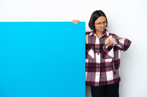 Jeune femme métisse avec une grande pancarte bleue isolée sur fond blanc montrant le pouce vers le bas avec une expression négative