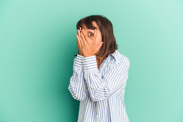 Jeune femme métisse isolée sur un clignotement bleu à travers les doigts effrayés et nerveux.