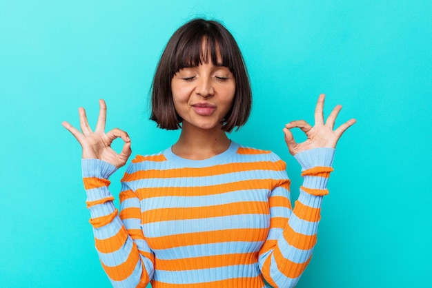 Jeune femme métisse isolée sur fond bleu se détend après une dure journée de travail, elle fait du yoga.