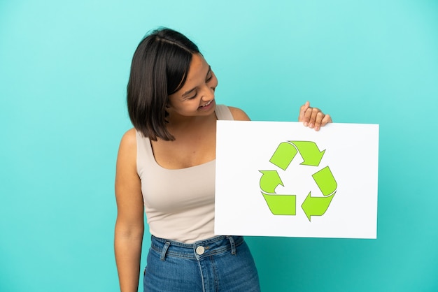 Jeune femme métisse isolée sur fond bleu tenant une pancarte avec l'icône de recyclage avec une expression heureuse