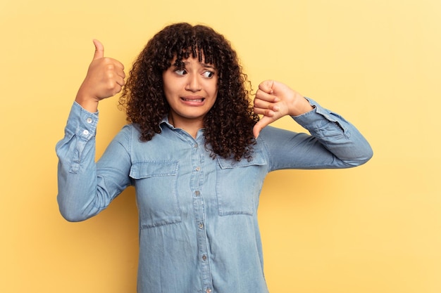 Jeune femme métisse isolée sur fond jaune montrant les pouces vers le haut et les pouces vers le bas difficile de choisir le concept